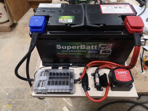 A car battery with red and blue terminals, mounted on a stand with wiring and fuses.