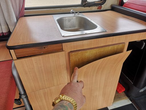 A hand reaches for a wooden cabinet door beside a small kitchen sink.
